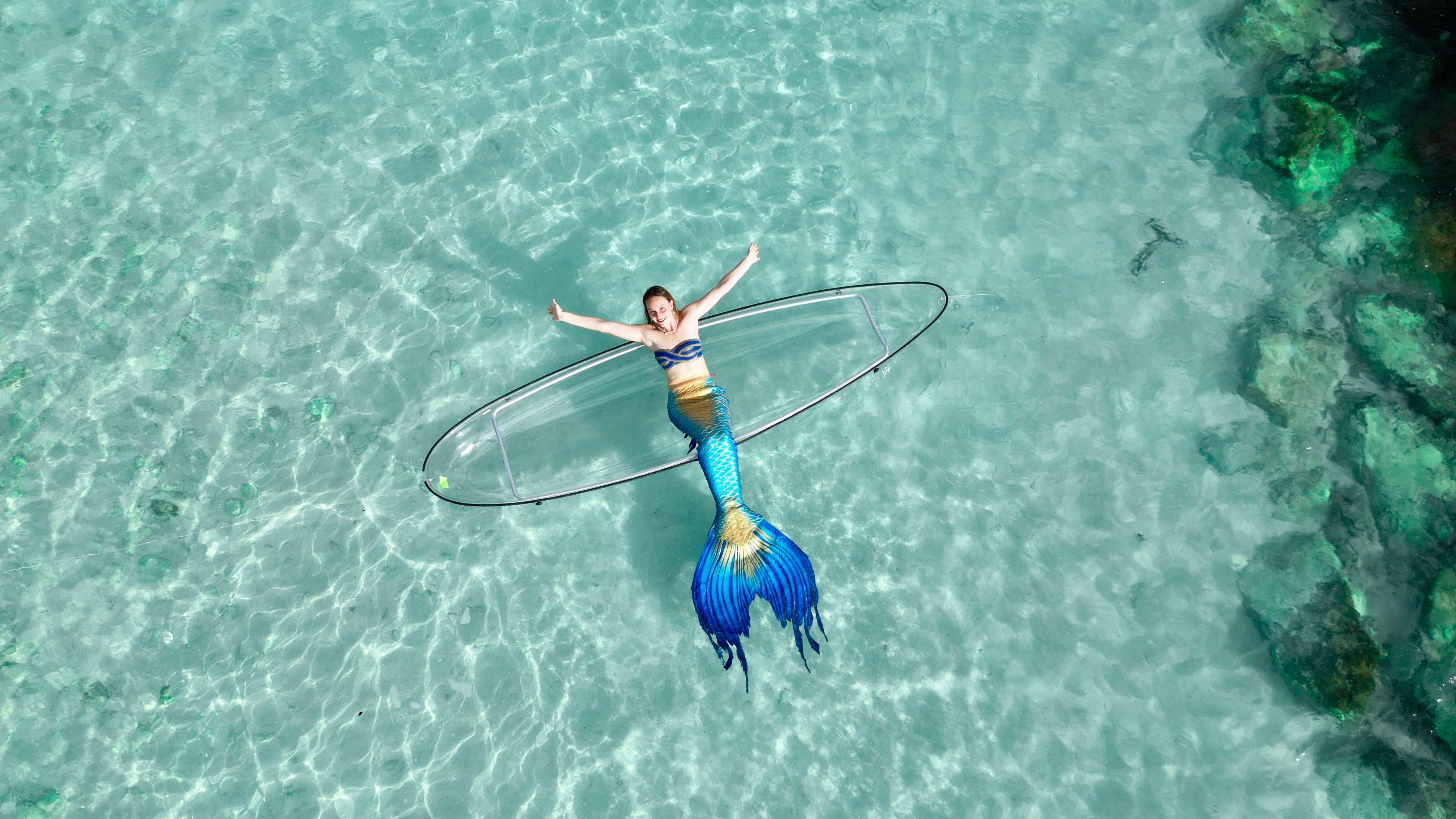 AquaMermaid founder Marielle in a blue silicone mermaid tail floating in turquoise water, reaching up toward a transparent kayak